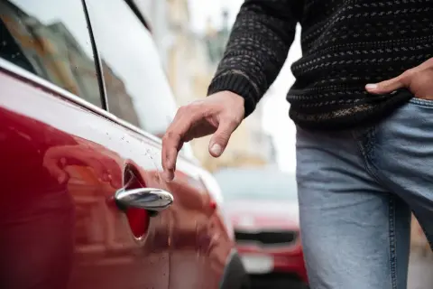 Mano tocando la manija de un vehículo rojo, con un suéter y jeans. Parece que el conductor está a punto de entrar.