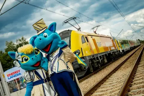 Dwa niebieskie smoki w strojach pozują przy żółtym pociągu. To specjalny pociąg do Energylandii, który zabierze Cię na niezapomnianą przygodę!