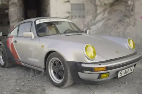 Srebrny Porsche 911 z żółtymi reflektorami, w stylu cyberpunk, stoi na tle zniszczonej ściany z graffiti.