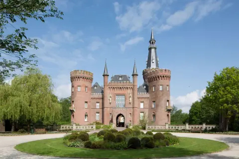 Schloss Moyland, ein imposantes Backsteinschloss mit Türmen und einem hohen Spitzdach, thront über einer gepflegten Gartenanlage mit einem runden Blumenbeet.