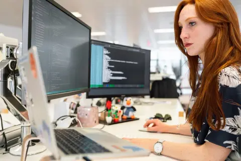 Rudowłosa programistka pracuje przy komputerze, monitor wyświetla kod. Czas na aktualizację sterowników.