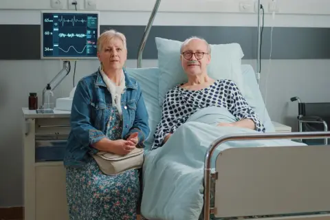 Starsza kobieta siedzi obok starszego mężczyzny w łóżku szpitalnym, na monitorze widoczne są parametry życiowe.