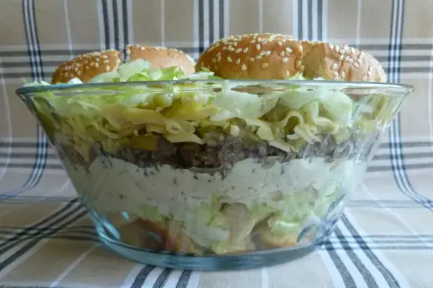 Warstwowa sałatka big mac przepis: mięso, ser, sałata, ogórek i sos w szklanej misie, zwieńczona bułkami z sezamem.
