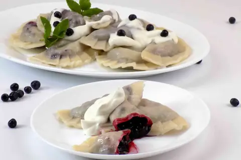 Pyszne pierogi z jagodami, polane śmietaną i ozdobione miętą. Na talerzu widać też kilka świeżych jagód.