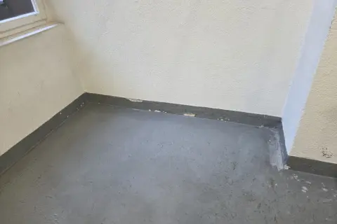 Ecke eines Raumes mit grauem Boden und weißer Wand. Am Sockel ist ein dunkler Streifen zu sehen, auf dem sich schimmel zieht von unten nach oben.
