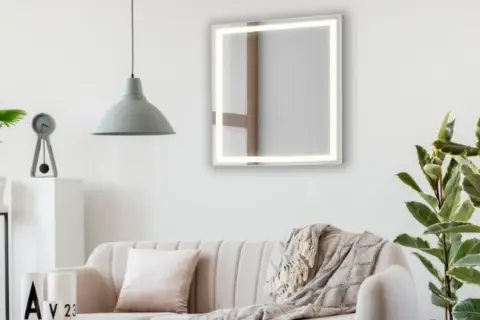 Un espejo iluminado sobre un sofá beige, con plantas y un reloj. Ideas de donde colocar espejos en el salón para ampliar el espacio.