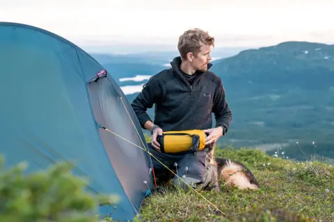 Man med hund förbereder tältet för en vandring i Norrland. Vacker fjällvy i bakgrunden.