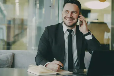 Uśmiechnięty mikroprzedsiębiorca w garniturze rozmawia przez telefon, notując coś w zeszycie. Obok leży laptop.