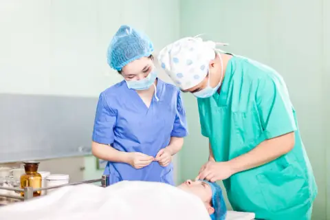 Deux chirurgiens masqués et coiffés préparent une intervention. L'un tient des instruments, l'autre s'occupe du patient. Ils se demandent "que veut dire DAE" dans ce contexte médical.