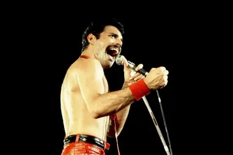 Freddie Mercury w czerwonych spodniach i opasce na nadgarstku śpiewa do mikrofonu. Nawet jako dorosły, jego energia przypominała dzikie dziecięce zabawy.
