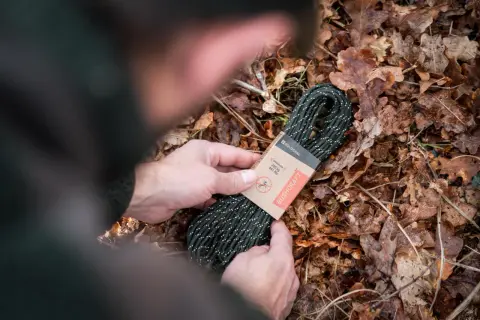 Manos sostienen un rollo de cordón paracord verde con puntos blancos, sobre un lecho de hojas secas.
