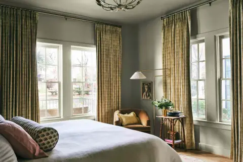 A cozy bedroom with a bed, a chair, and a floor lamp. The curtains are a beautiful millenial green, adding a touch of nature to the room.
