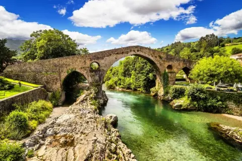 Majestuoso puente romano asturias sobre un río de aguas cristalinas, rodeado de exuberante vegetación y un cielo azul con nubes blancas.