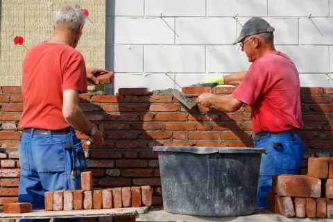Dwóch budowlańców stosuje zasady murowania z cegły pełnej, układając cegły na zaprawie.