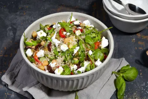 Bunter Quinoa Salat mit Kichererbsen, Feta, Oliven und Rucola, serviert in einer Schüssel. Daneben ein Glas mit Kräuter Dressing.