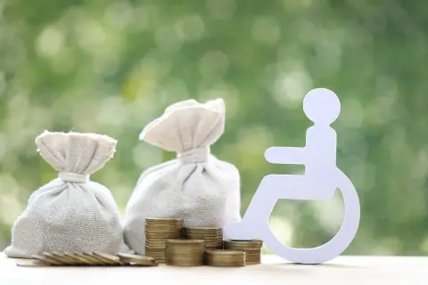 Symbol osoby na wózku inwalidzkim obok worków z pieniędzmi i monet sugeruje świadczenie wspierające kwoty.