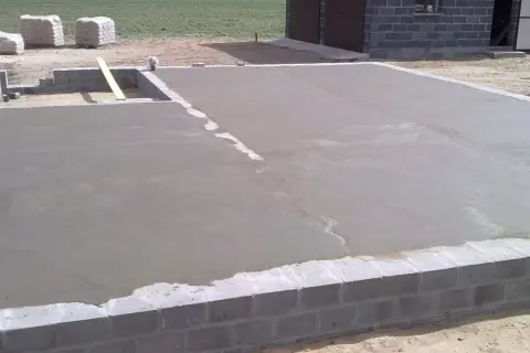 Świeżo wylany chudy beton na fundamencie domu. W tle widać materiały budowlane i fragment budynku.