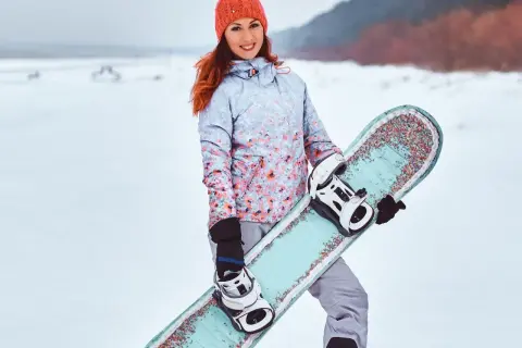 Uśmiechnięta dziewczyna w czapce i kurtce z kwiatowym wzorem trzyma **co to jest snowboard** z kolorowymi zdobieniami.
