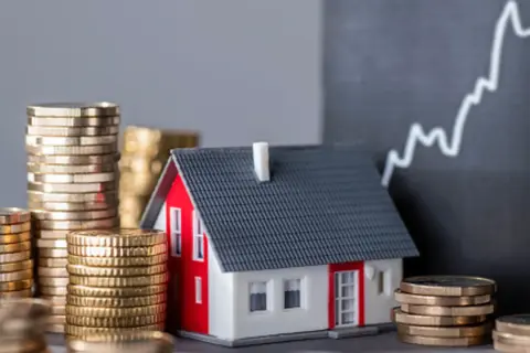 Casa modelo rodeada de pilas de monedas, con un gráfico ascendente al fondo, sugiriendo un estudio de solvencia y el valor de la inversión inmobiliaria.