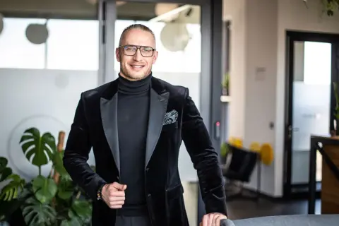 Mężczyzna w czarnym garniturze i okularach stoi w nowoczesnym biurze.