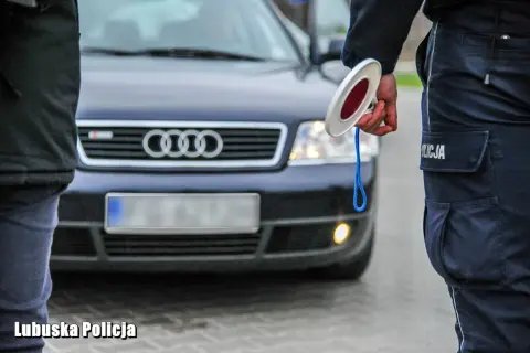 Policjant z lizakiem zatrzymuje auto. Widać jego mundur i fragment ciemnego Audi. Jakie światła podczas deszczu, gdy policja kontroluje ruch.