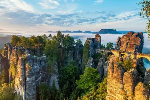 Felsformationen und eine Basteibrücke in der Böhmischen Schweiz, umgeben von Nebel und Bäumen, unter einem blauen Himmel.