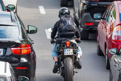 Motocyklista w kasku i plecaku przeciska się między samochodami w korku. Widać znaki motocyklistów na tablicy rejestracyjnej.