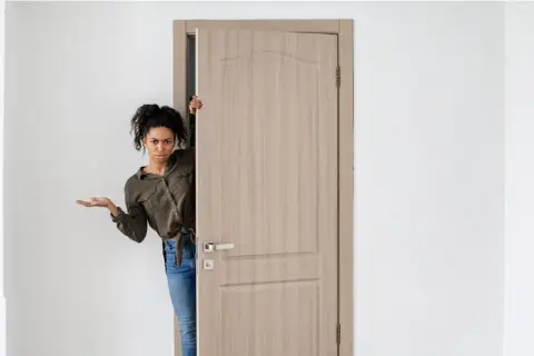 Mujer asomándose por una puerta de madera clara, con expresión de duda. Los colores de puertas son neutros y elegantes.