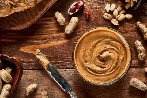 Cremige Erdnussbutter in einer Schüssel, daneben Erdnüsse und ein Messer. Erdnussbutter gesund und lecker!