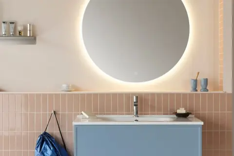 Ett modernt badrum med en stor, upplyst rund spegel som ger en känsla av rymd. Att inreda med speglar är smart! Blått kommod och rosa kakel skapar en mjuk atmosfär.