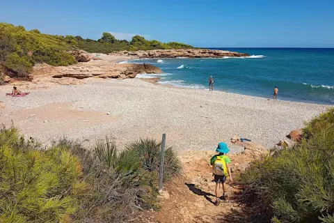 Niño con mochila azul desciende por sendero hacia la playa de rueta, con rocas y mar turquesa.