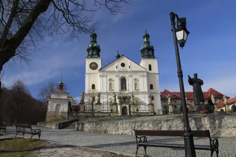 Kalwaria Zebrzydowska: barokowy kościół z wieżami, plac z ławkami i pomnik.