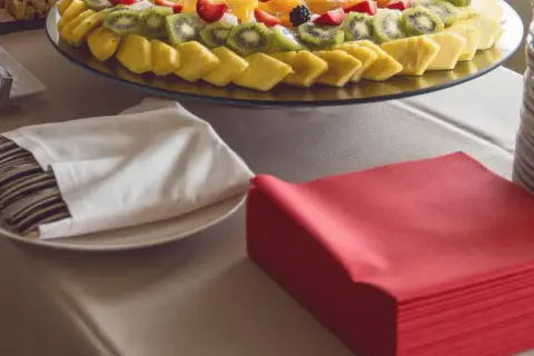 Une table de restaurant prête pour un repas, avec un plateau de fruits frais, des couverts emballés dans des serviettes blanches et des serviettes rouges pliées.