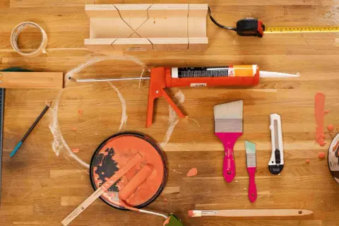 Un tableau peinture acrylique sur bois, prêt pour un projet de bricolage. Pinceaux, rouleau, pot de peinture orange, pistolet à mastic, ruban adhésif, mètre et équerre sont disposés sur un plan de travail en bois.
