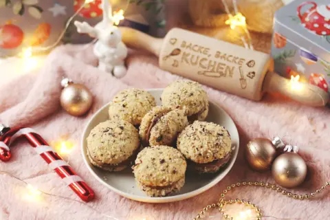 Leckere Nutella Kekse, umhüllt von gehackten Nüssen, liegen auf einem Teller. Ein Nudelholz mit der Aufschrift "Backe, backe Kuchen" und Weihnachtsdeko sorgen für festliche Stimmung.