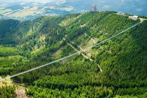 Najdłuższy most w Czechach, Sky Bridge 721, przecina zieloną dolinę pełną drzew. Na szczycie góry widać wieżę widokową.