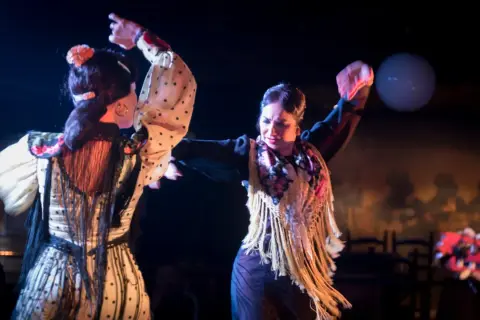 Dos bailarinas de flamenco, con trajes de lunares y flecos, interpretan apasionadas canciones de sevillanas bajo luces de escenario.