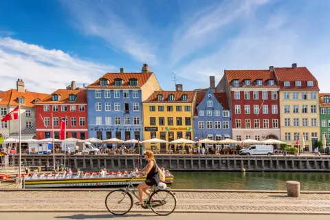 En kvinna cyklar längs Nyhavn i Köpenhamn, ett soligt exempel på danmark klimat. Färgglada hus och kanalbåtar skapar en livlig atmosfär.