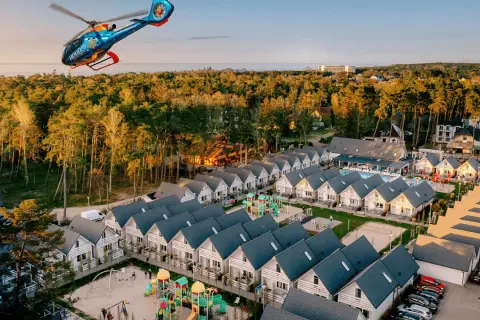 Widok z lotu ptaka na holiday park z domkami, placem zabaw i basenem, a nad nim latający helikopter.