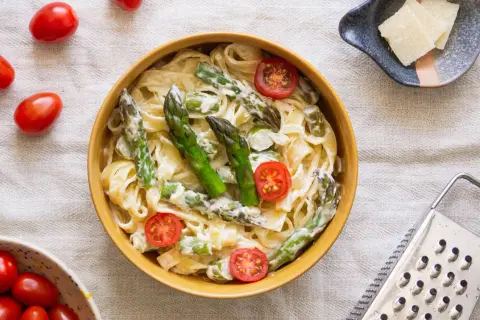 Pyszny makaron ze szparagami w sosie śmietanowym, udekorowany pomidorkami koktajlowymi. Obok miseczka z parmezanem i tarką.