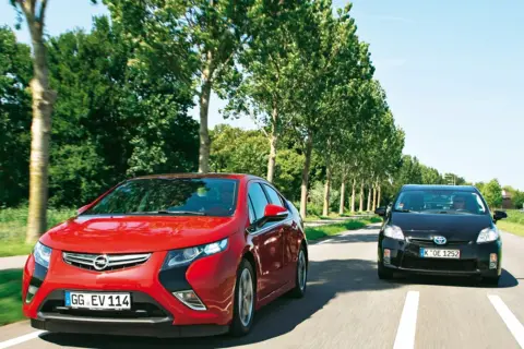 Czerwona Opel Ampera i czarny samochód jadą drogą otoczoną drzewami.