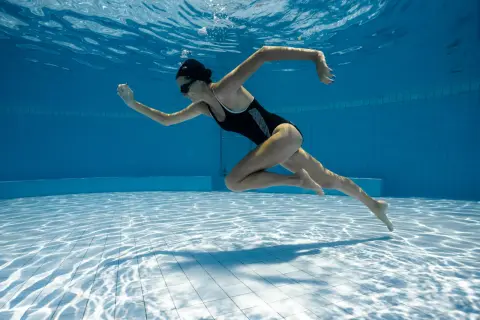 Underwater shot of a woman in a black swimsuit performing aqua aerobics. Her body is streamlined as she propels herself through the blue water.