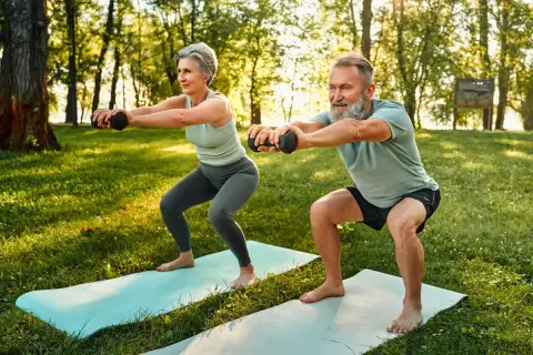 Une femme sportive de 50 ans et un homme font des squats avec des haltères dans un parc ensoleillé.