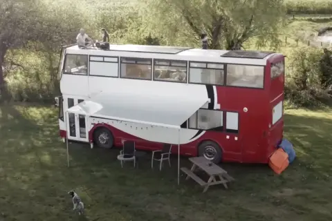 Autobús motorhome de dos pisos con paneles solares en el techo. Personas sentadas en la parte superior, un toldo extendido y muebles de exterior.