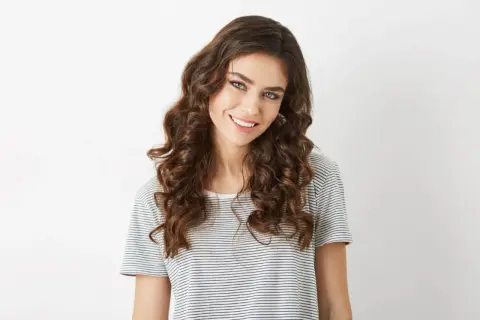 Mujer sonriente con pelo ondulado castaño, ojos azules y camiseta a rayas.