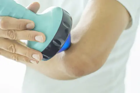 Une main utilise un appareil bleu clair pour soigner une tendinite au coude. La lumière bleue émise par l'appareil cible la zone douloureuse.