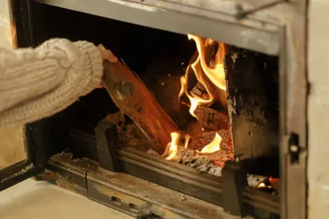 Main tenant ajoutant une bûche dans un poêle à bois, les flammes dansent joyeusement.