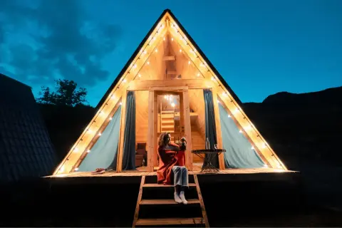 Soirée de camping nature corse. Une femme assise sur les marches d'un chalet en bois illuminé par des guirlandes lumineuses, profitant de la nuit.
