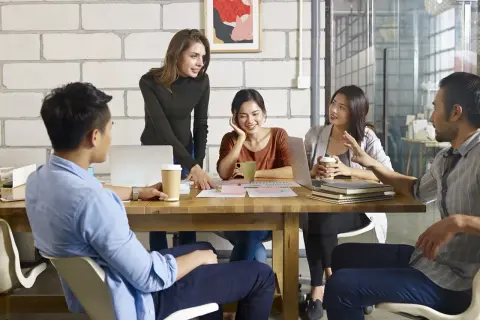 Team discute progetti, con una donna che indica documenti e un uomo che parla. Si discute di trasferte in busta paga.