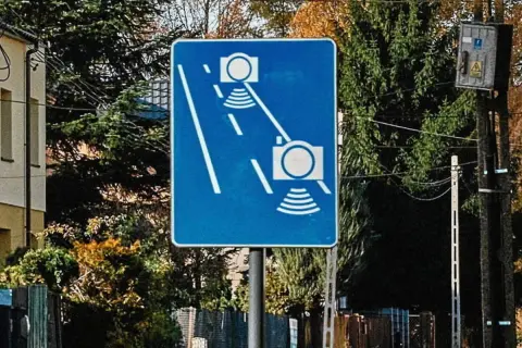 Niebieski znak drogowy z białymi symbolami aparatów fotograficznych i falami, informujący o odcinkowym pomiarze prędkości.